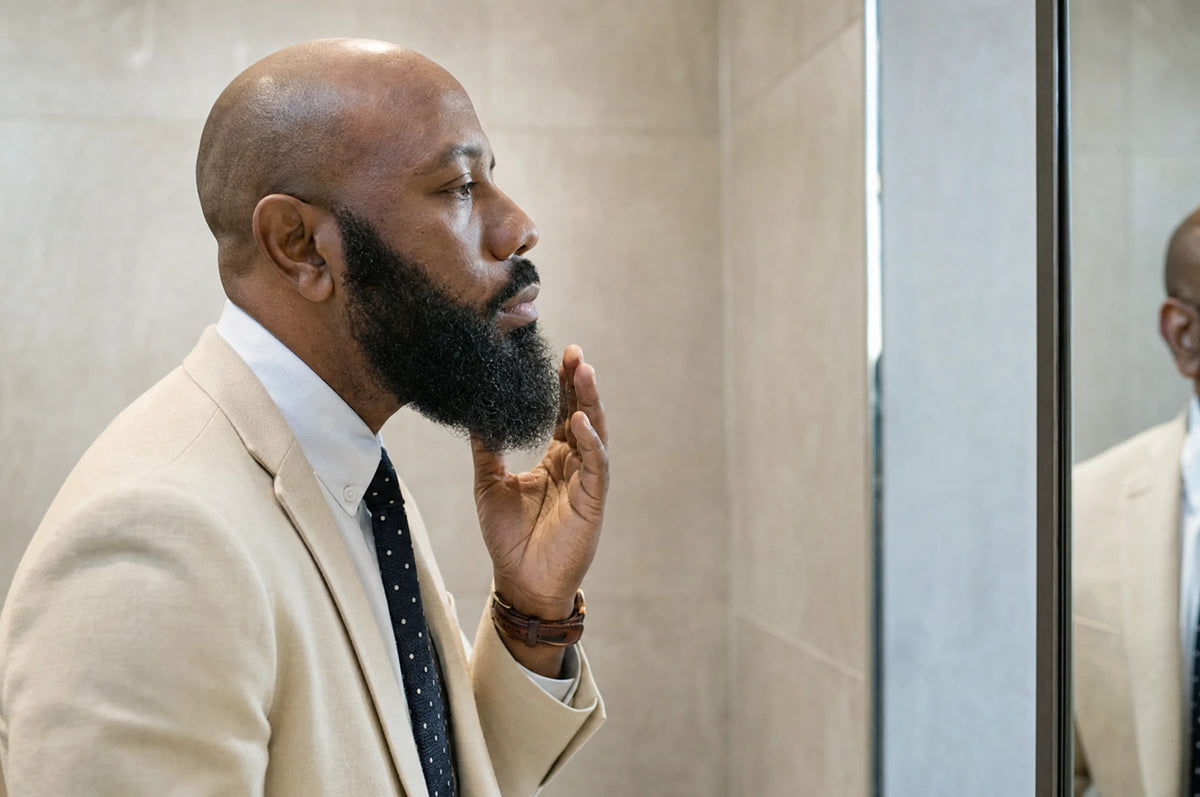 Homme chauve en costume crème observant sa barbe fournie dans un miroir