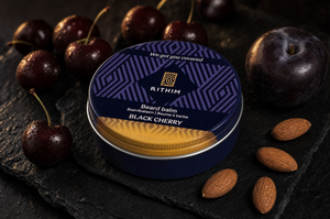 A round tin of Rithim Beard Balm in Black Cherry scent, styled on a dark slate with fresh black cherries, a plum, and almonds.